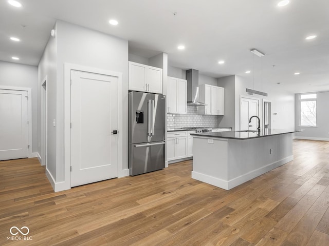 kitchen with white cabinets, stainless steel fridge with ice dispenser, dark countertops, light wood-type flooring, and wall chimney range hood