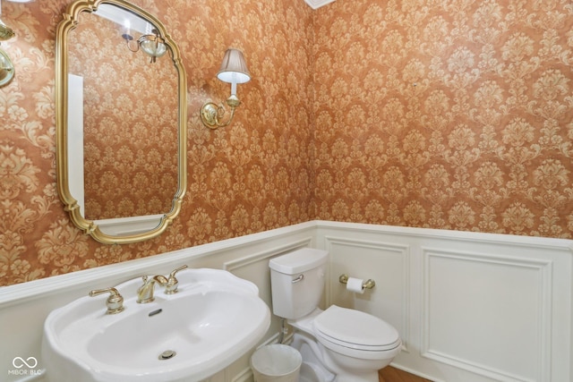half bath with a sink, wallpapered walls, a wainscoted wall, and toilet