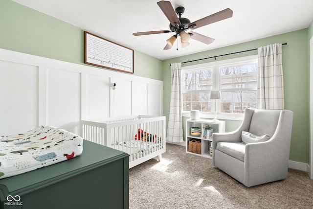 carpeted bedroom featuring a decorative wall, wainscoting, a ceiling fan, and a nursery area