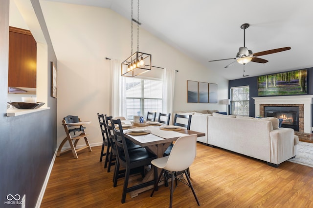 dining room featuring baseboards, ceiling fan with notable chandelier, a fireplace, light wood-style floors, and high vaulted ceiling