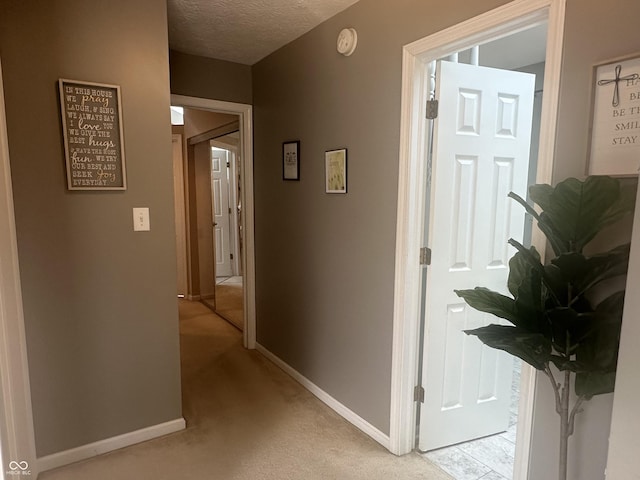 hall featuring a textured ceiling, light colored carpet, and baseboards