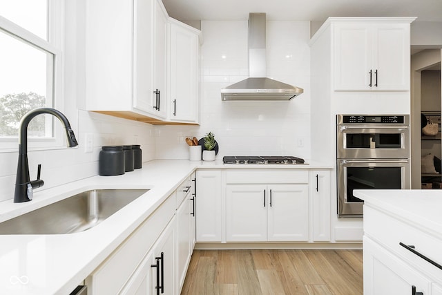 kitchen with white cabinets, wall chimney exhaust hood, double oven, gas stovetop, and a sink