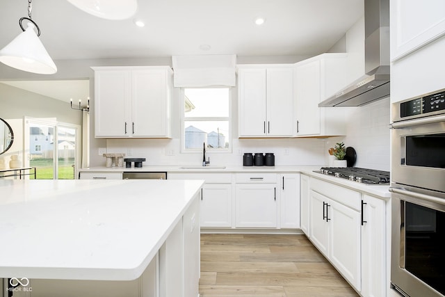 kitchen with appliances with stainless steel finishes, light countertops, light wood-type flooring, wall chimney range hood, and a sink