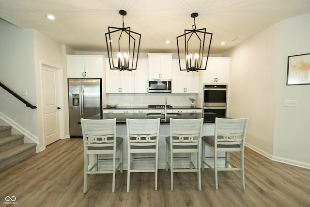 kitchen with dark countertops, decorative backsplash, appliances with stainless steel finishes, a kitchen island with sink, and a chandelier