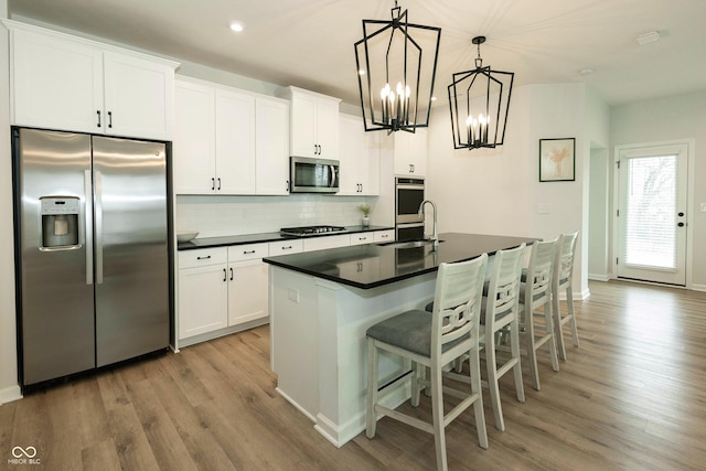 kitchen featuring tasteful backsplash, dark countertops, appliances with stainless steel finishes, light wood-style floors, and an island with sink