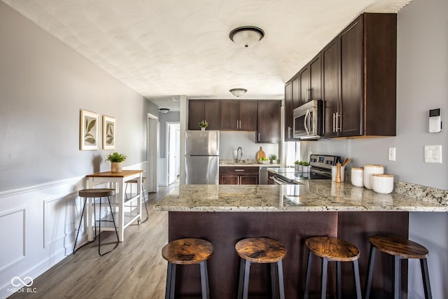 kitchen with dark brown cabinetry, appliances with stainless steel finishes, a peninsula, light wood-style floors, and a sink