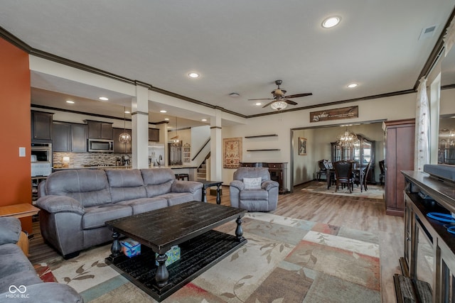 living area with ceiling fan with notable chandelier, crown molding, light wood-style flooring, and stairs