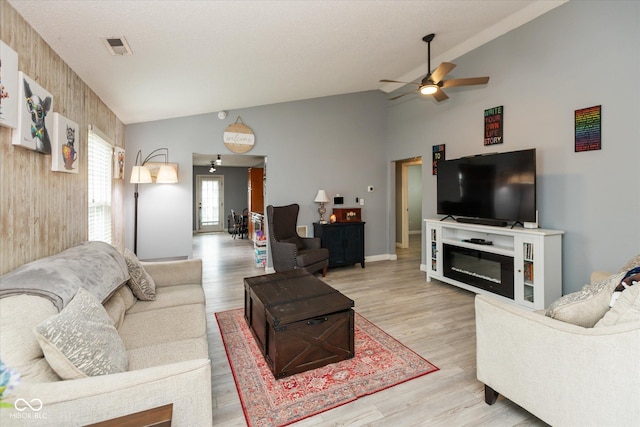 living room featuring lofted ceiling, ceiling fan, visible vents, light wood finished floors, and a glass covered fireplace