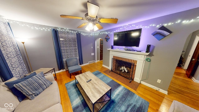 living area featuring arched walkways, a ceiling fan, a brick fireplace, wood finished floors, and baseboards