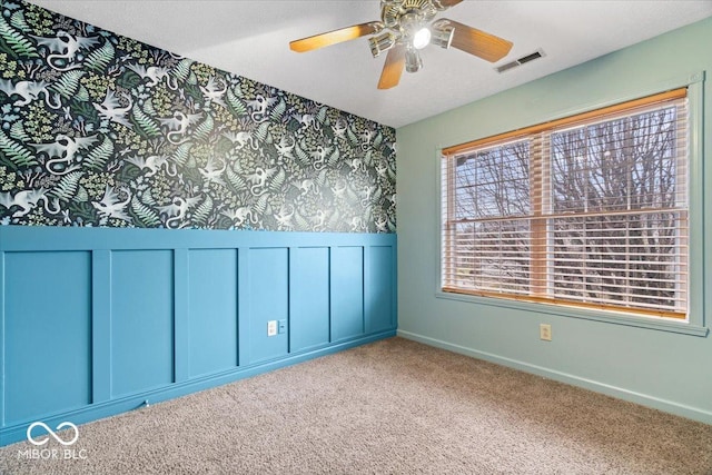 carpeted spare room with a wainscoted wall, baseboards, visible vents, wallpapered walls, and ceiling fan