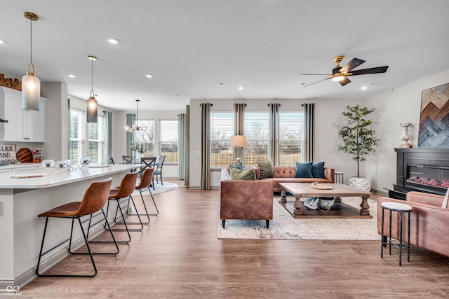 living area with ceiling fan with notable chandelier, a glass covered fireplace, recessed lighting, light wood-style floors, and baseboards