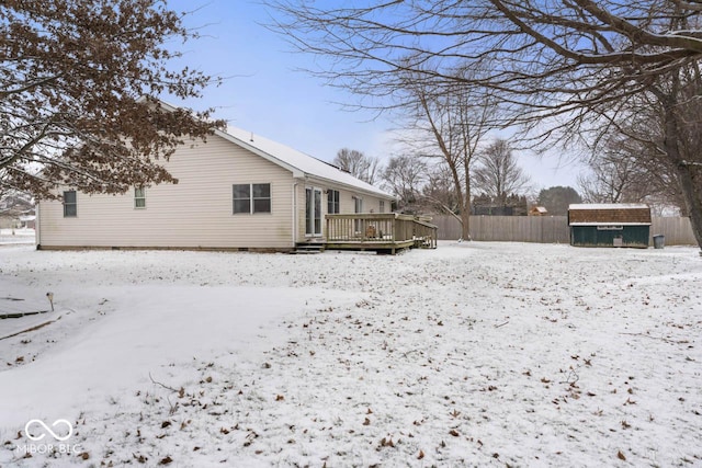 snow covered house with a wooden deck, a storage shed, an outdoor structure, and fence