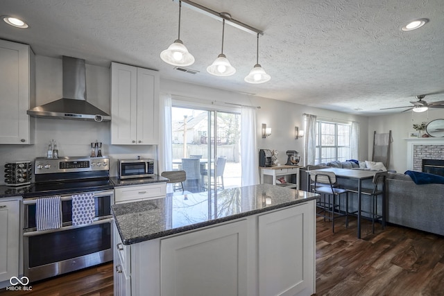 kitchen with wall chimney range hood, dark wood-style flooring, appliances with stainless steel finishes, a brick fireplace, and open floor plan