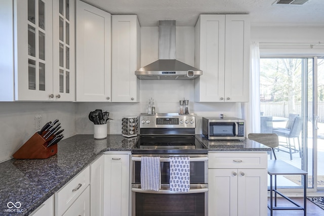 kitchen with glass insert cabinets, dark stone counters, appliances with stainless steel finishes, white cabinetry, and wall chimney exhaust hood