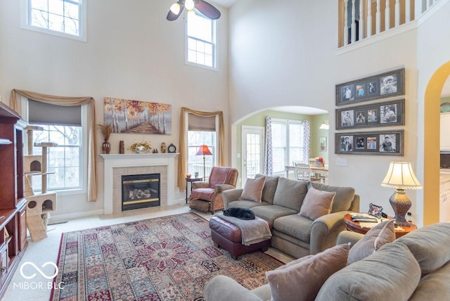 living room featuring arched walkways, a ceiling fan, a tile fireplace, a towering ceiling, and carpet