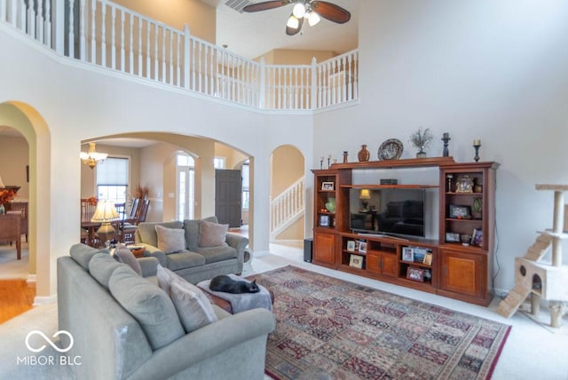 carpeted living area with stairway, a high ceiling, arched walkways, and ceiling fan with notable chandelier