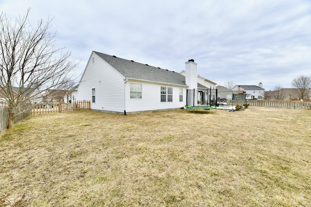 rear view of property with a trampoline, roof with shingles, a yard, a chimney, and a fenced backyard