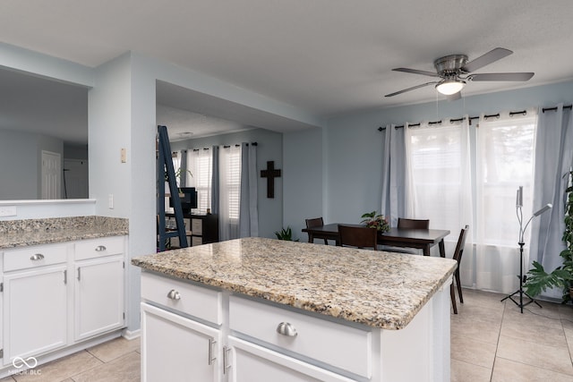 kitchen with light tile patterned flooring, a kitchen island, a ceiling fan, white cabinets, and light stone countertops