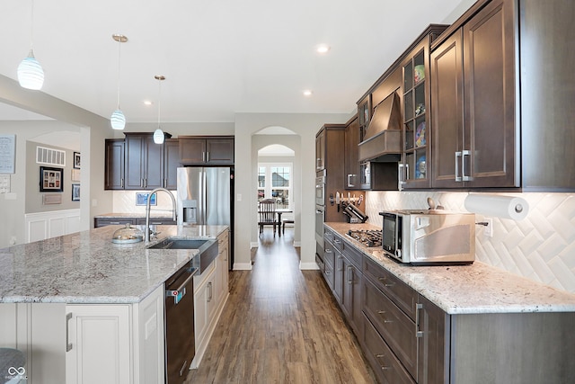 kitchen with arched walkways, custom range hood, appliances with stainless steel finishes, dark brown cabinetry, and a sink