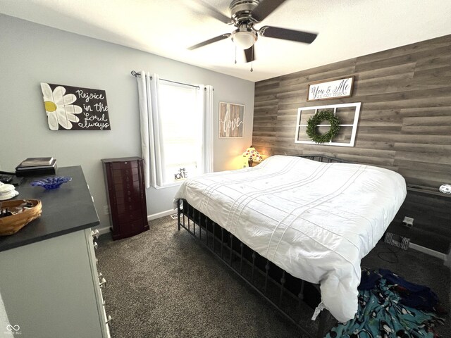 bedroom with a ceiling fan, wooden walls, dark colored carpet, baseboards, and an accent wall