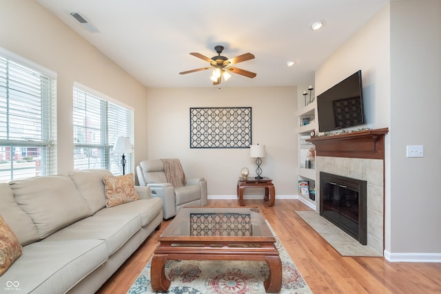 living area featuring baseboards, visible vents, a ceiling fan, a tiled fireplace, and light wood-style flooring