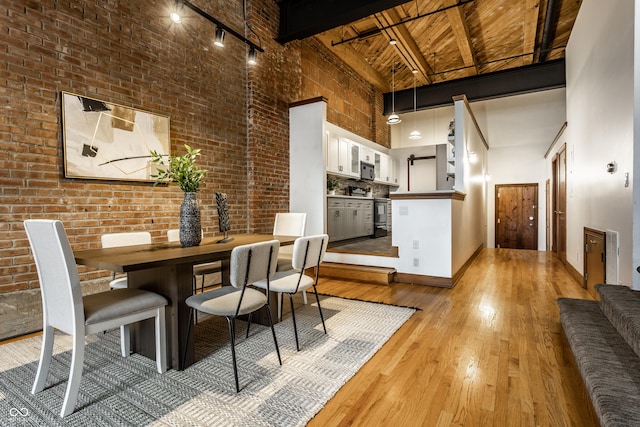 dining room featuring light wood finished floors, wooden ceiling, brick wall, beamed ceiling, and a high ceiling