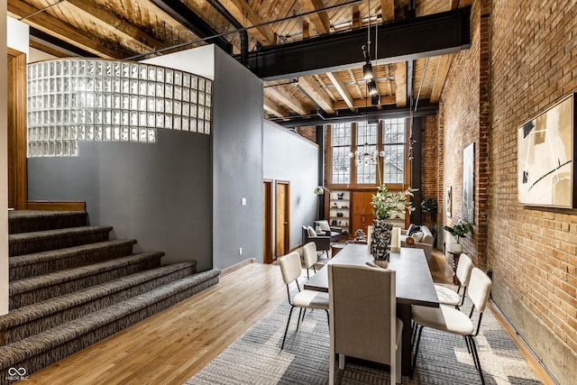 dining space featuring brick wall, a high ceiling, wood finished floors, beam ceiling, and an inviting chandelier