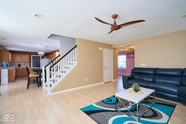 living area with ceiling fan with notable chandelier, stairway, light wood-style flooring, and baseboards