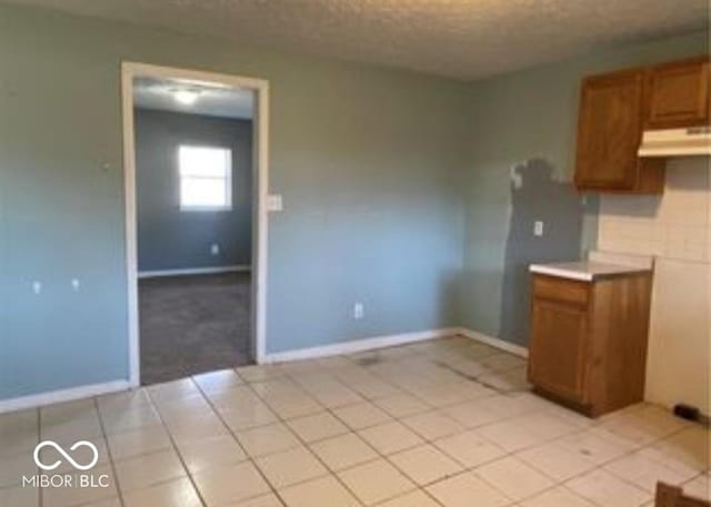 kitchen featuring brown cabinets, light tile patterned flooring, a textured ceiling, under cabinet range hood, and baseboards