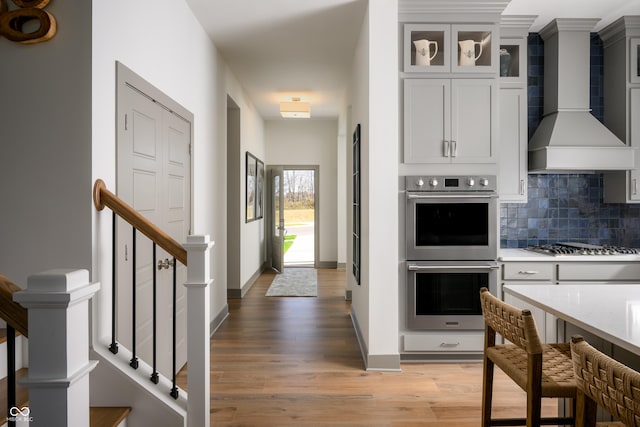 kitchen with appliances with stainless steel finishes, backsplash, wall chimney exhaust hood, light wood finished floors, and glass insert cabinets