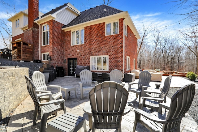 rear view of house featuring brick siding, an outdoor living space, a shingled roof, a chimney, and a patio