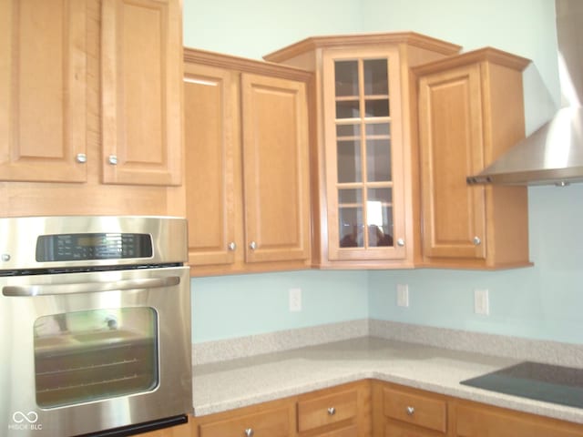 kitchen featuring wall chimney exhaust hood, glass insert cabinets, black electric stovetop, light countertops, and stainless steel oven