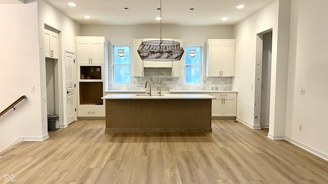 kitchen with a kitchen island with sink, a sink, white cabinets, light wood-style floors, and tasteful backsplash