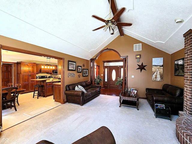 living room featuring a ceiling fan, visible vents, vaulted ceiling with beams, a textured ceiling, and light colored carpet