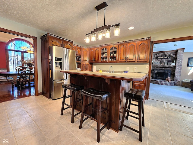 kitchen featuring a brick fireplace, glass insert cabinets, light countertops, a kitchen bar, and stainless steel refrigerator with ice dispenser