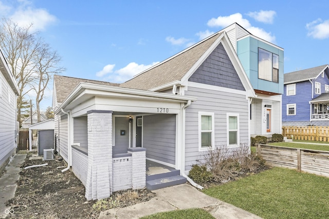 view of front of home featuring a shingled roof, covered porch, central AC unit, fence, and a front lawn