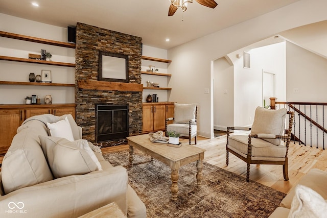 living room featuring light wood-type flooring, visible vents, a ceiling fan, arched walkways, and a stone fireplace