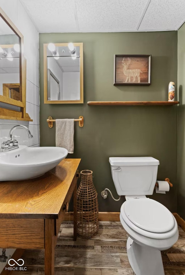 half bathroom with toilet, wood finished floors, a paneled ceiling, baseboards, and vanity