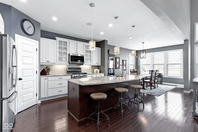 kitchen with dark wood-style floors, a kitchen island with sink, a sink, glass insert cabinets, and appliances with stainless steel finishes