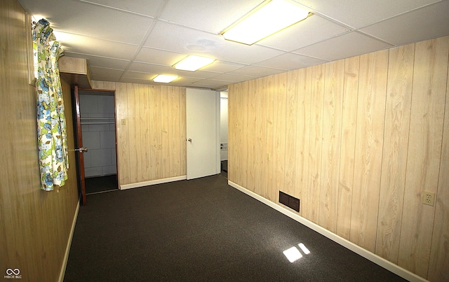 empty room with a paneled ceiling, wood walls, carpet flooring, visible vents, and baseboards