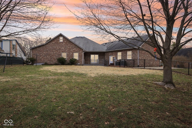 back of house at dusk with roof with shingles, brick siding, a lawn, and fence