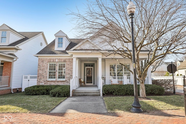 view of front facade with a front lawn, fence, covered porch, a shingled roof, and brick siding