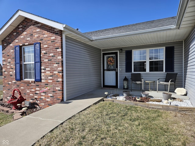 doorway to property featuring covered porch, brick siding, and roof with shingles