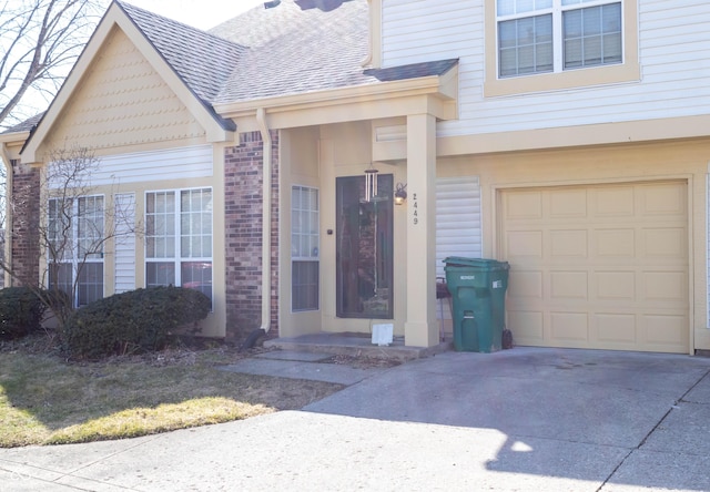 entrance to property featuring brick siding, an attached garage, a shingled roof, and driveway