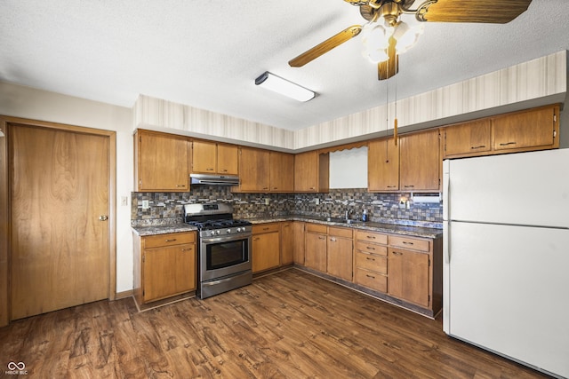 kitchen featuring under cabinet range hood, dark wood-style floors, freestanding refrigerator, brown cabinetry, and stainless steel gas range