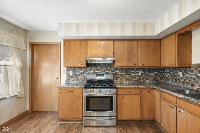 kitchen with wood finished floors, wallpapered walls, under cabinet range hood, a textured ceiling, and gas range