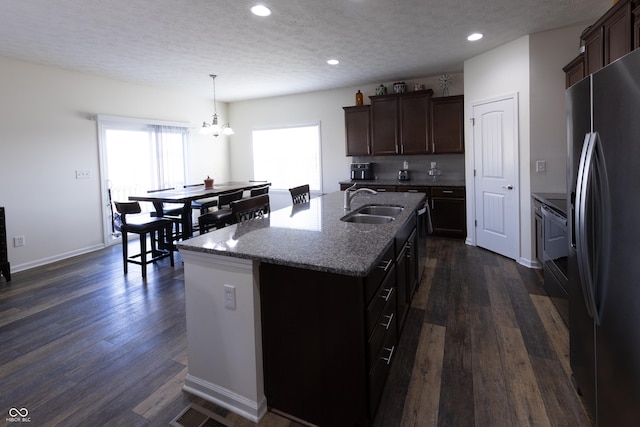 kitchen with a sink, an island with sink, stainless steel refrigerator with ice dispenser, stone countertops, and dark wood-style flooring