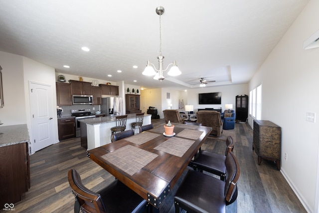 dining space with baseboards, dark wood finished floors, recessed lighting, a raised ceiling, and ceiling fan with notable chandelier