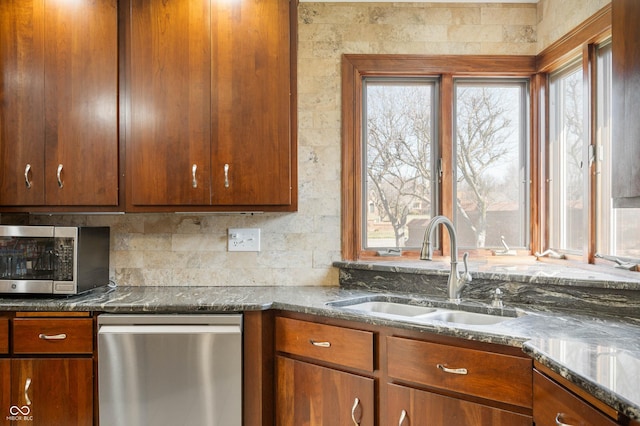 kitchen with brown cabinetry, dark stone counters, stainless steel appliances, a sink, and tasteful backsplash