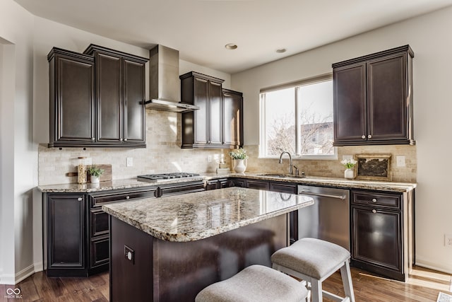 kitchen featuring a kitchen island, a sink, dark wood-type flooring, appliances with stainless steel finishes, and wall chimney range hood